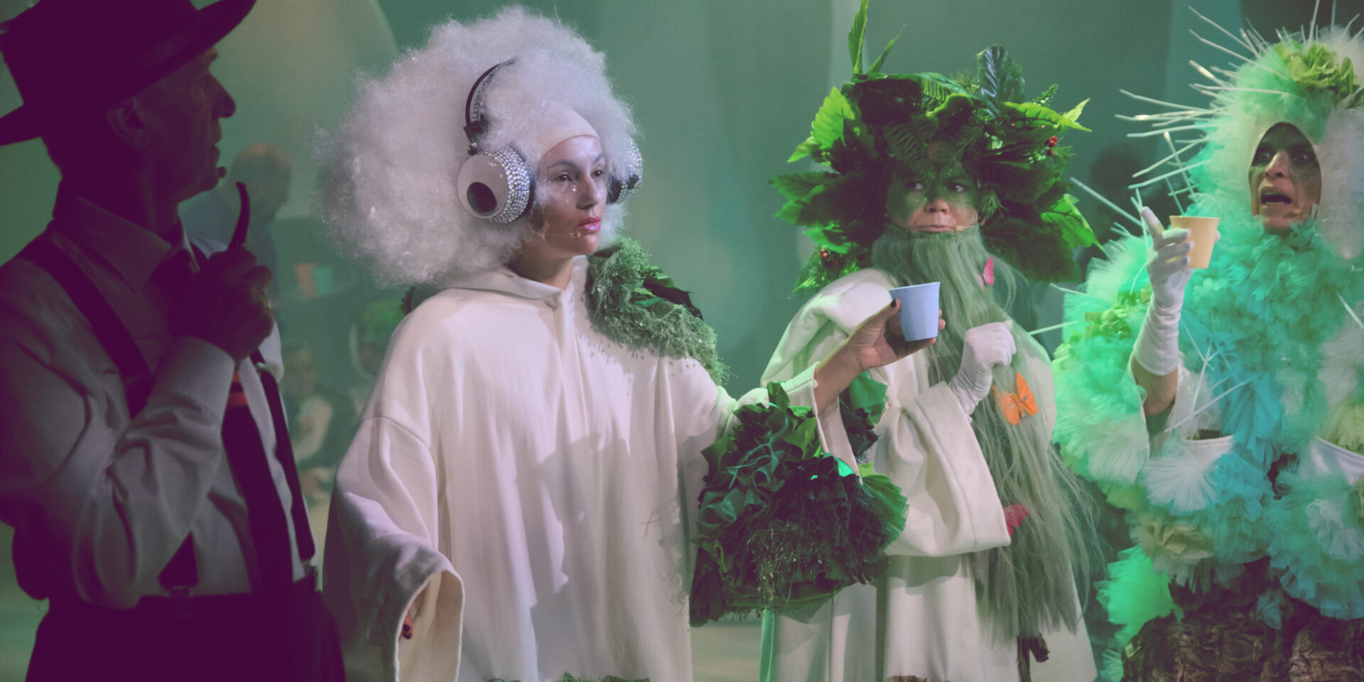 Cztery postacie ustawione w rzędzie. Po lewej mężczyzna w kapeluszu z fajką, obok niego kobieta ubrana na biało w słuchawkach na głowie i z kubkiem w dłoni. Po prawej dwie postaci ubrane w biało zielone stroje ozdobione elementami przypominającymi liście i mech.