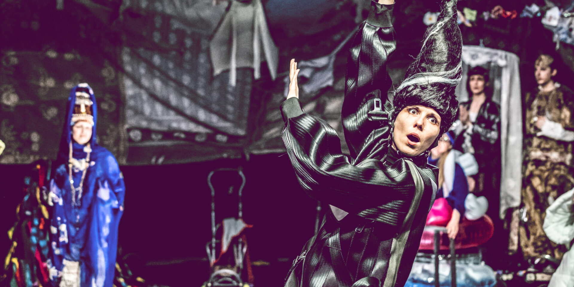 Na środku stoi kobieta w czarnej peruce w kształcie stożka. Ma uniesione do góry ręce.