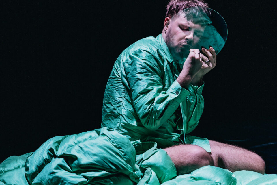 Mężczyzna w zielonej koszuli siedzi, przysłania do ust okrągły kawałek szkła, jedną ręką trzyma szkło, drugą dotyka ust w tym miejscu gdzie ma usta.